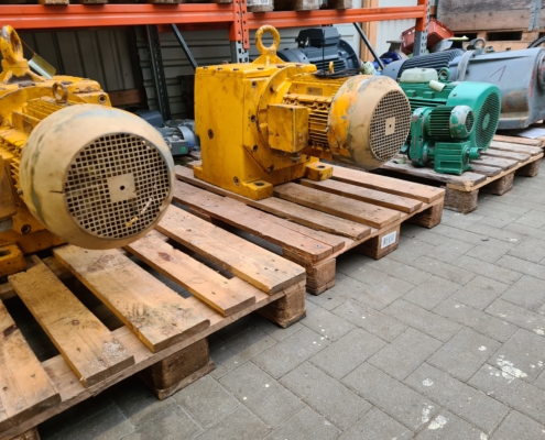 Mehrere Elektromotoren in Gelb und Grün auf Holzpaletten in einem Lager mit Regalen im Hintergrund