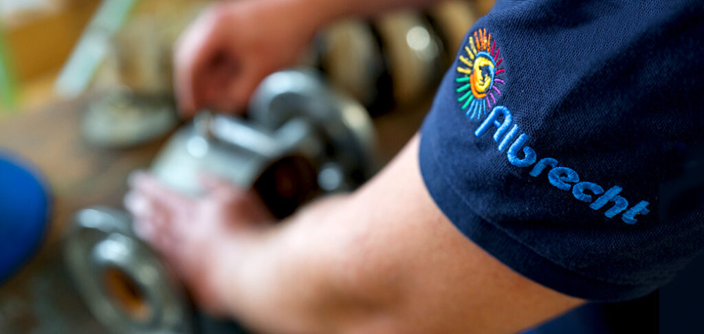 Arm mit blauem Shirt mit Aufschrift 'Albrecht' und farbigem Logo, der an einem Metallteil arbeitet