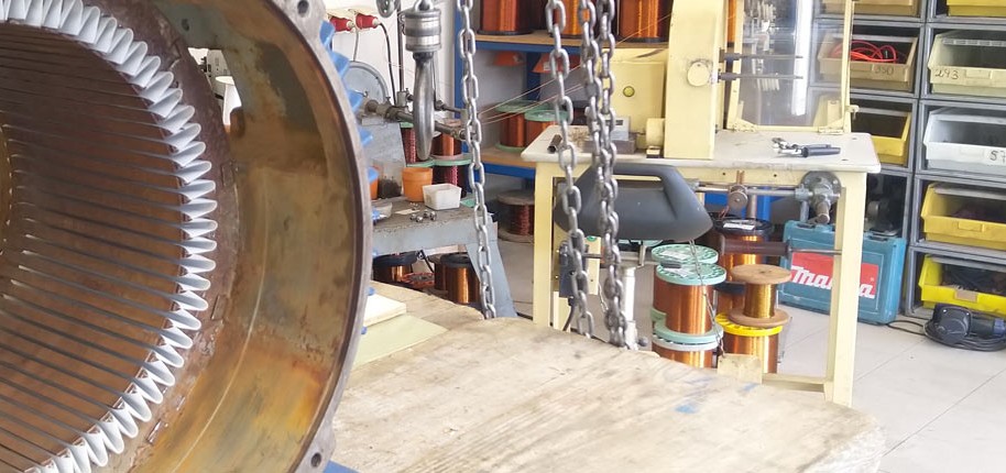 Werkstatt mit großem Elektromotor-Stator auf einem Holztisch, Ketten zum Heben und Regale mit Werkzeugen im Hintergrund