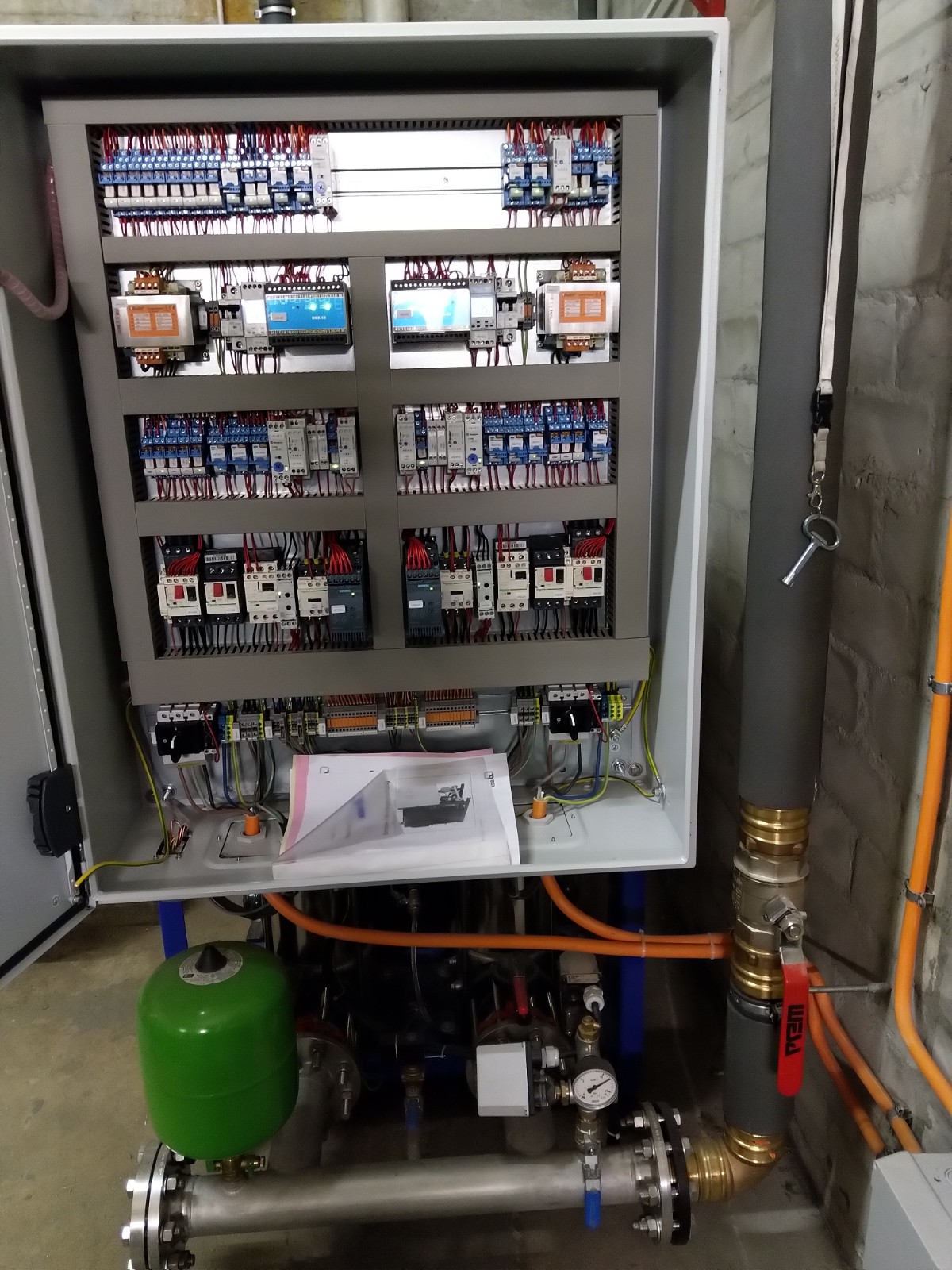 Geöffneter Schaltschrank mit zahlreichen elektrischen Komponenten und Kabeln, darunter Relais und Sicherungen, darunter Rohrleitungen mit Ventil und Manometer