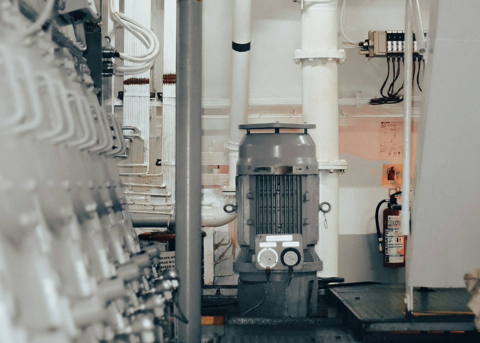 Industrieller Maschinenraum mit grauer Maschine in der Mitte, weißen Rohren und Feuerlöscher an der Wand