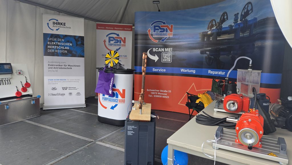 Messestand bei der MeLa 2025 mit zwei großen Bannern, einem gelben Ventilator, einer Kupferrohrinstallation und einer roten Pumpe. Banner zeigen Logos und Text zu Dienstleistungen.