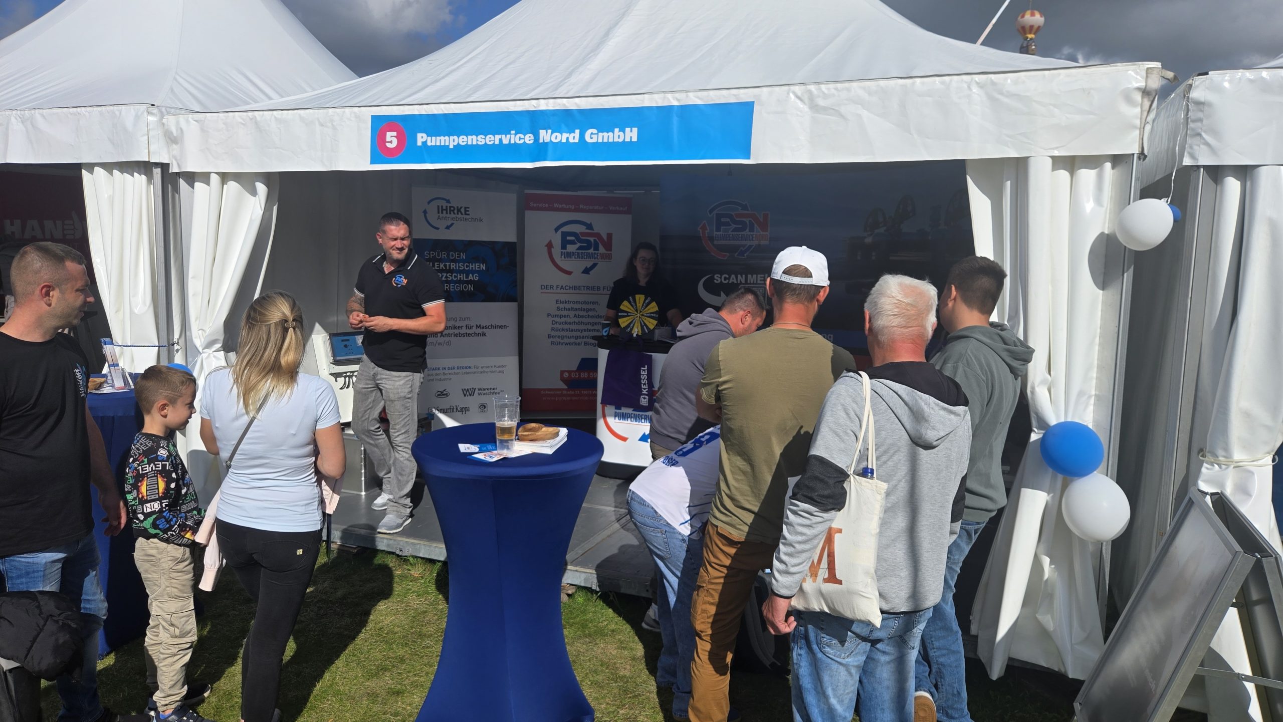Menschen stehen in einer Reihe vor einem Stand mit blauer Tischdecke. Auf dem Stand sind Banner mit 'Pumpenservice Nord GmbH' und Logos sichtbar. Einige Personen halten Informationsmaterial.