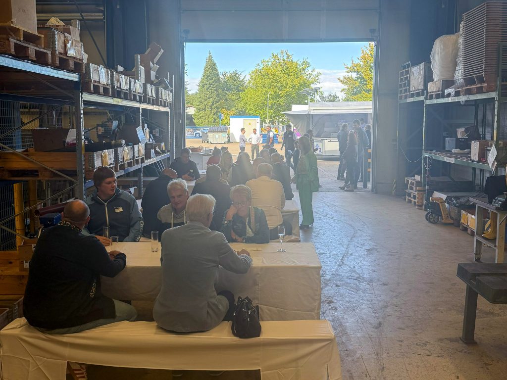 Mehrere Personen feiern das Sommerfest an langen Tischen mit weißen Tischdecken in einem Lagerraum , im Hintergrund ein offenes Tor mit Blick auf Bäume und Himmel.