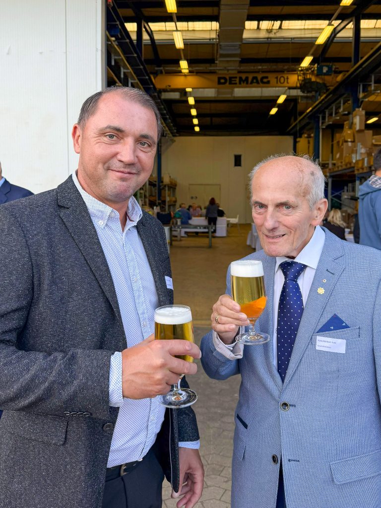 Templin und Kuni in Anzügen halten Gläser mit Bier, im Hintergrund eine Lagerhalle mit Regalen und einem Kran mit der Aufschrift 'DEMAG 10t'.