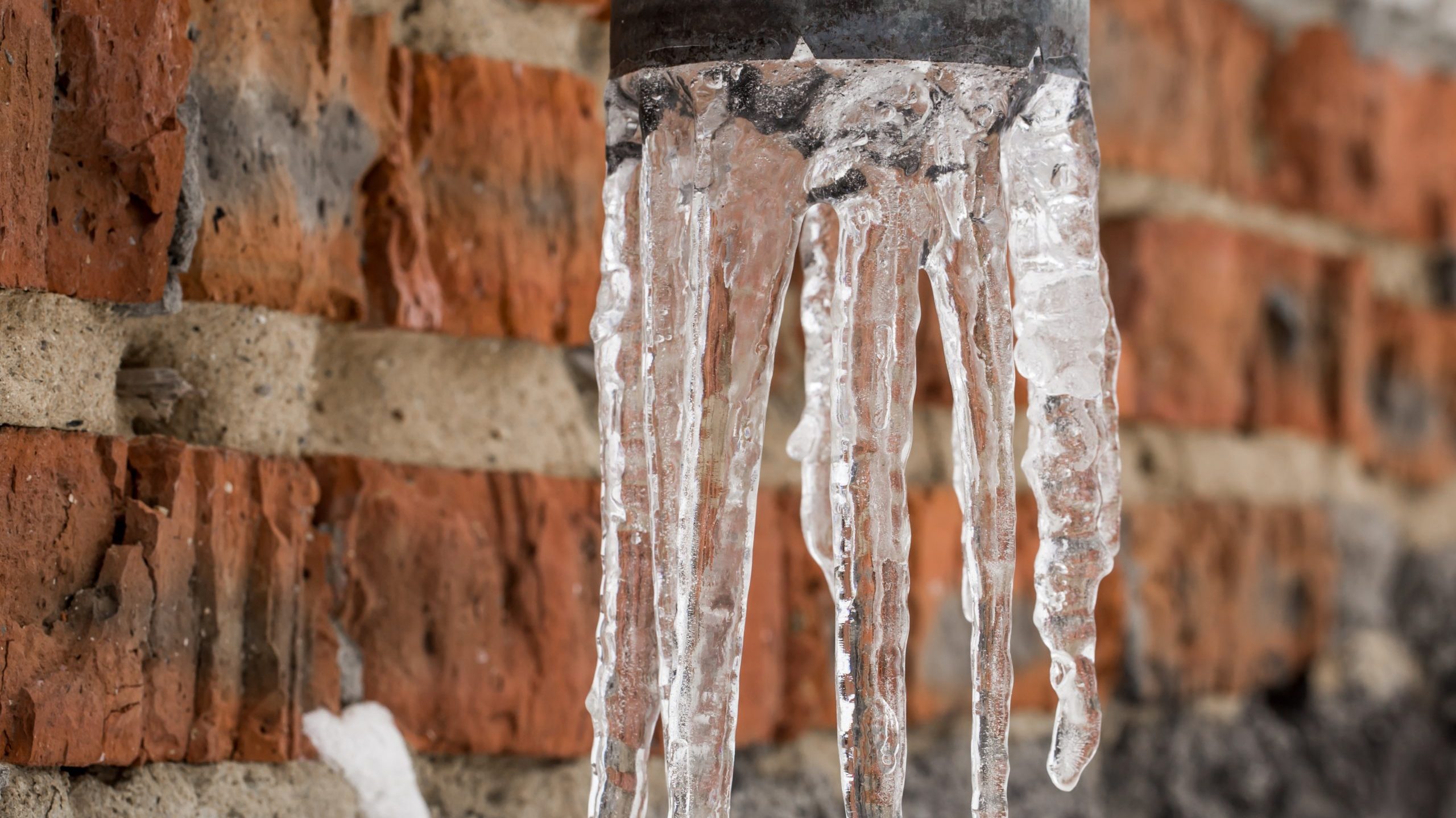Mehrere durchsichtige Eiszapfen hängen an einem nicht winterfestem grauen Metallrohr vor einer roten Backsteinmauer.