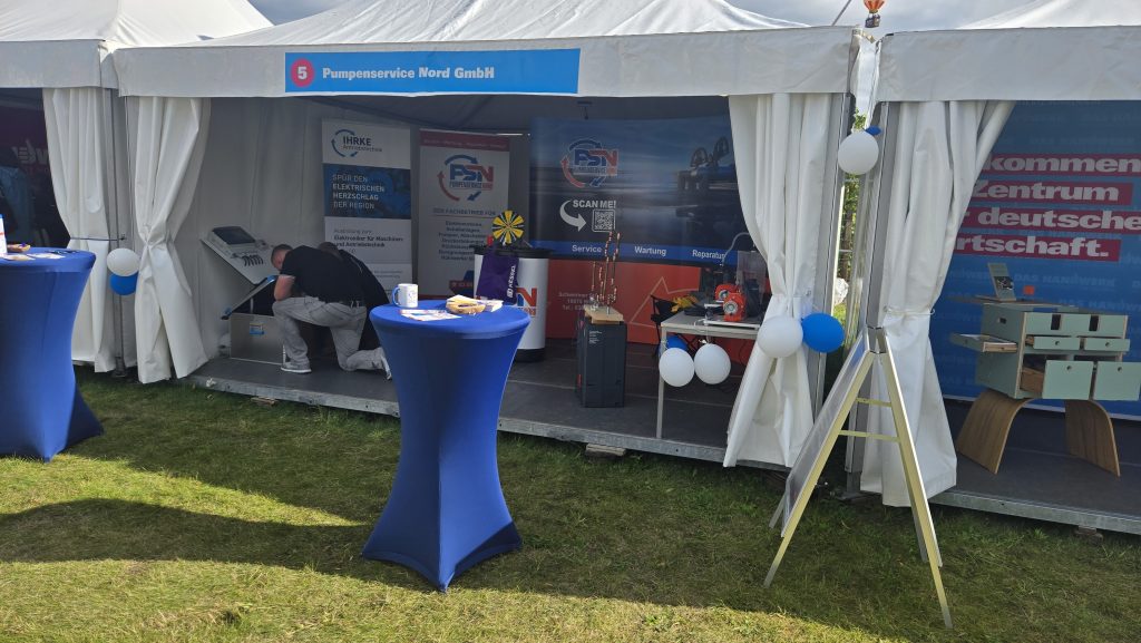 Zelt mit Informationsständen für die Mela 2025, blauer Stehtisch mit Snacks, Person bückt sich, Luftballons an Ständer und Zelt befestigt.