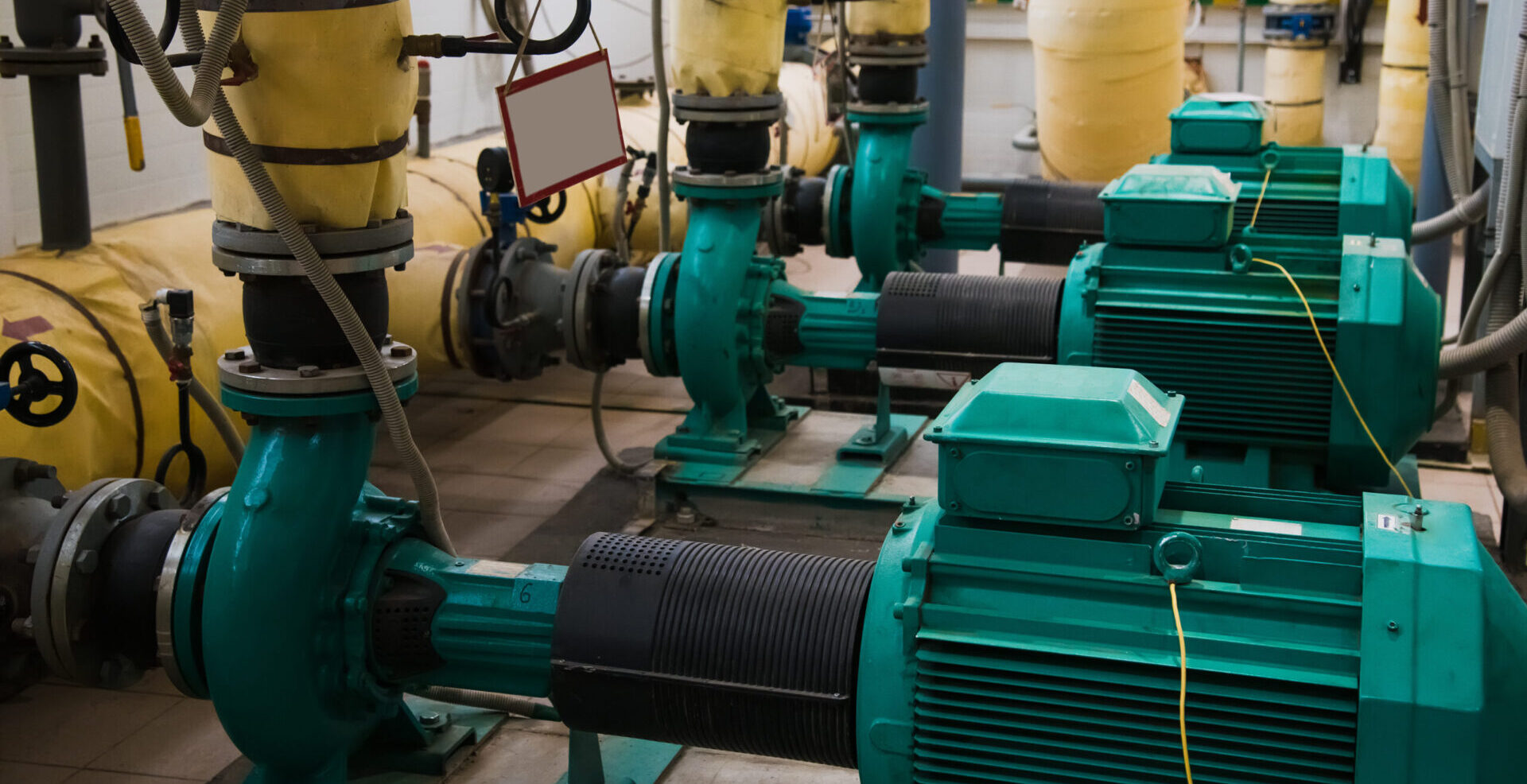 Nahaufnahme von mehreren Abwasserpumpen mit grünen Elektromotoren, gelben Rohrleitungen und sichtbaren Manometern sowie Ventilen.