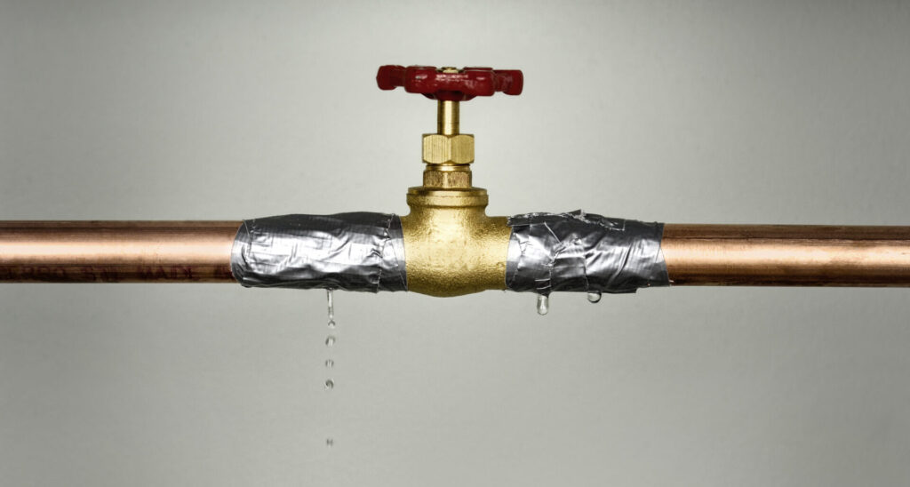 Nahaufnahme eines undichten Wasserrohrs mit rotem Ventil und austretenden Wassertropfen, Rohrverbindung mit silbernem Klebeband umwickelt. Das kann zu Trockenlauf führen.