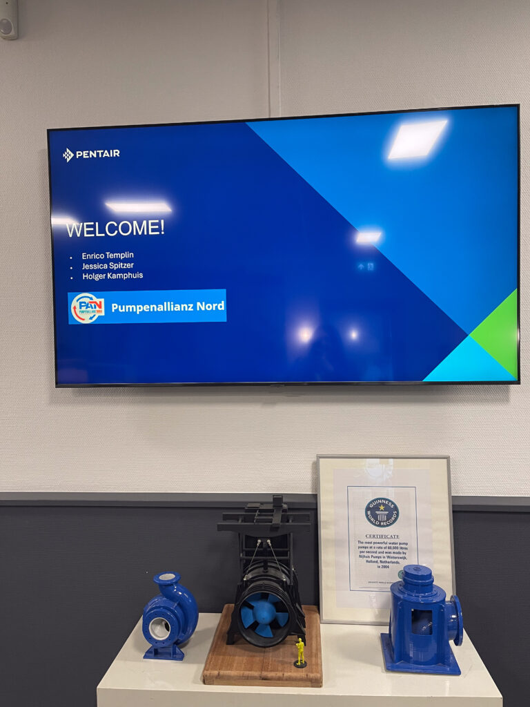 Pentair-Bildschirm mit Begrüßungstext, daneben zwei blaue Pumpenmodelle und eine gerahmte Urkunde auf einem Tisch