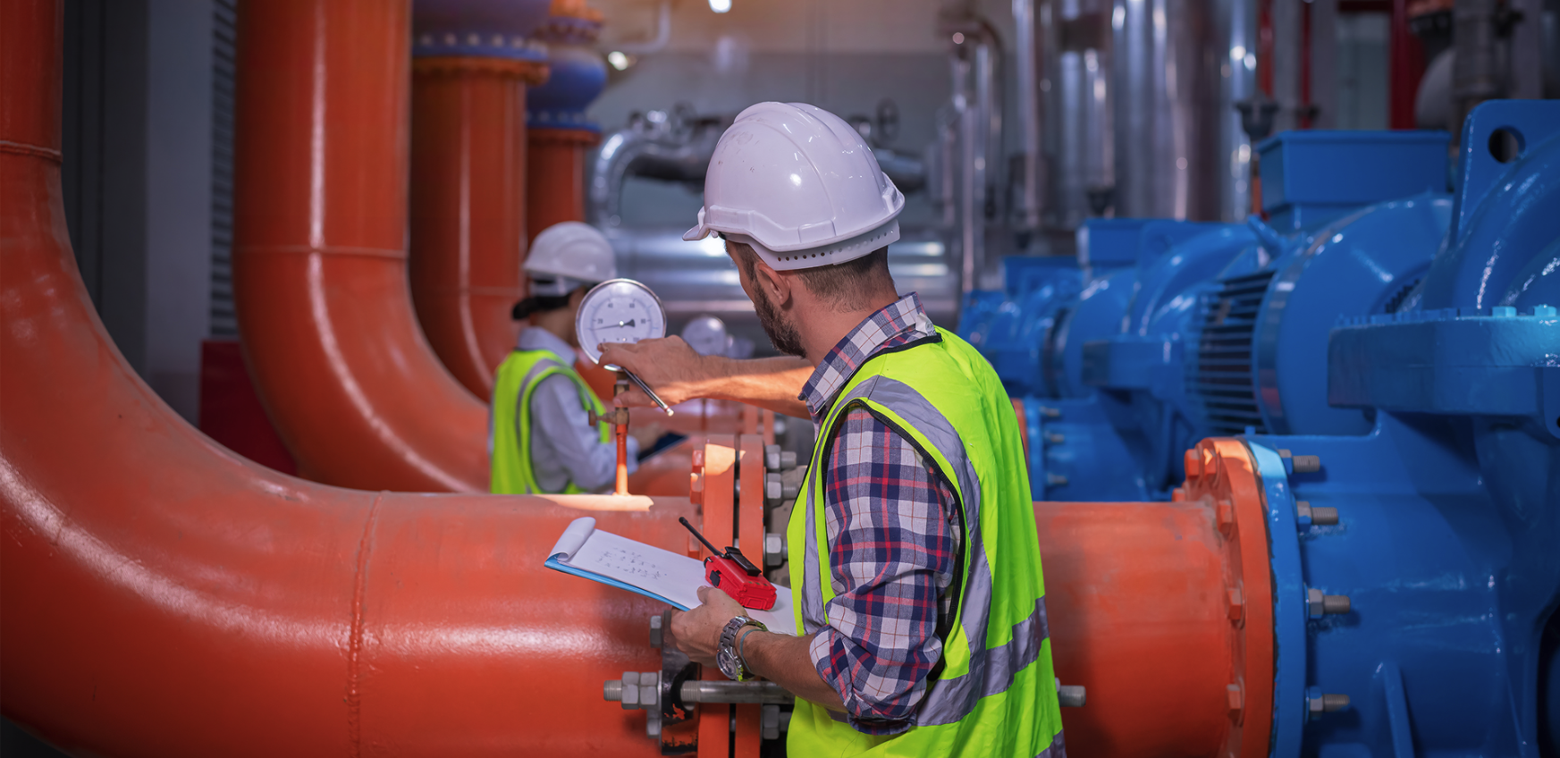 Ein Industrieingenieur mit Schutzhelm und Warnweste prüft mit einem Klemmbrett große orangefarbene Rohrleitungen in einer Fabrikhalle beim Montieren.