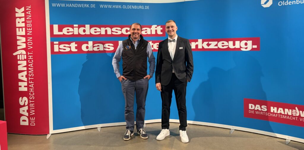 Zwei Männer stehen vor einer blauen Messewand mit mehreren roten Textbalken und Logos.