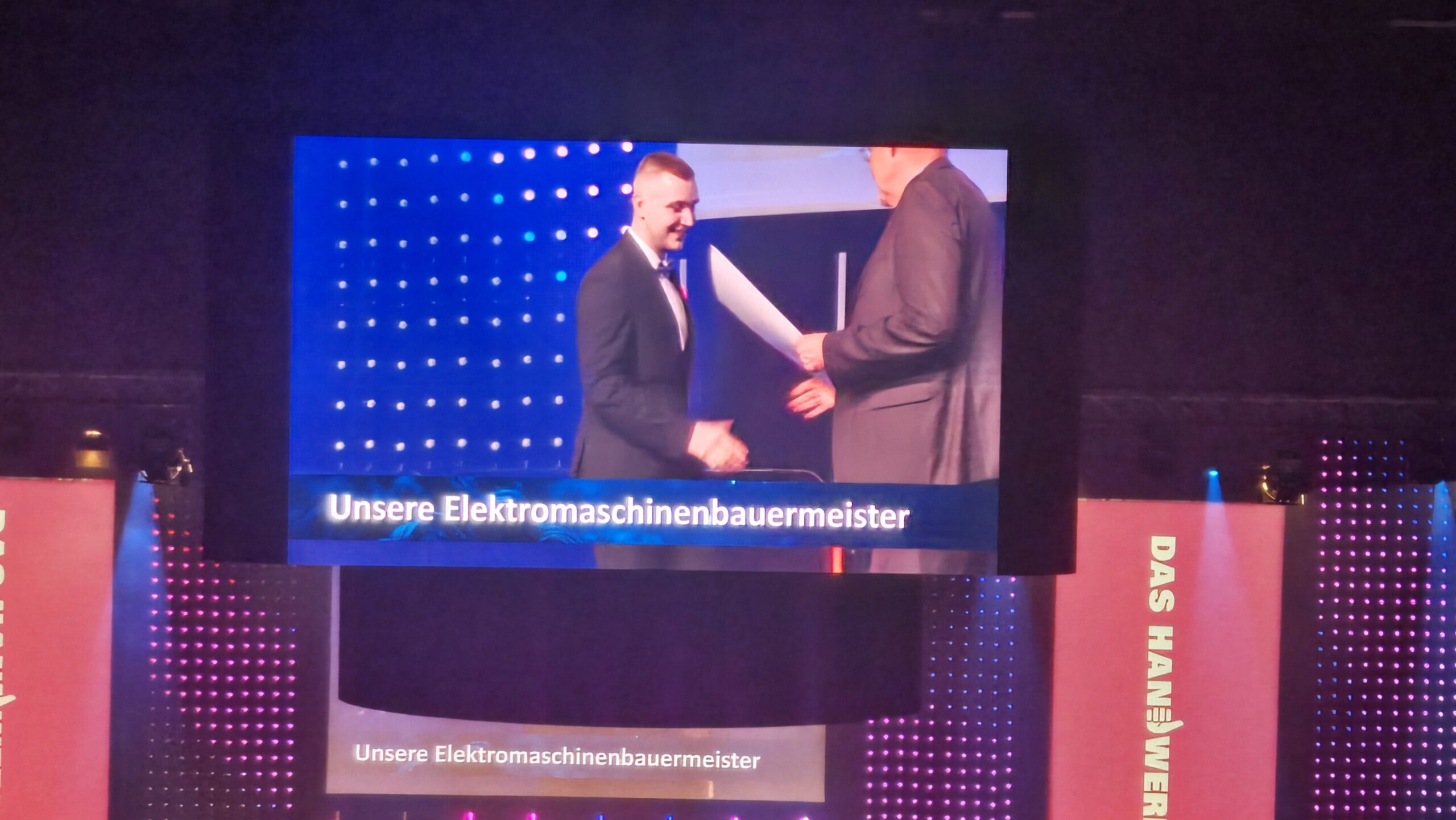 Kevin Bauer im Anzug auf einer Bühne bei der Meisterfeier 2026, während er von einer Person eine Urkunde überreicht bekommt, im Hintergrund große Leinwand mit der Aufschrift 'Unsere Elektromaschinenbauermeister'.