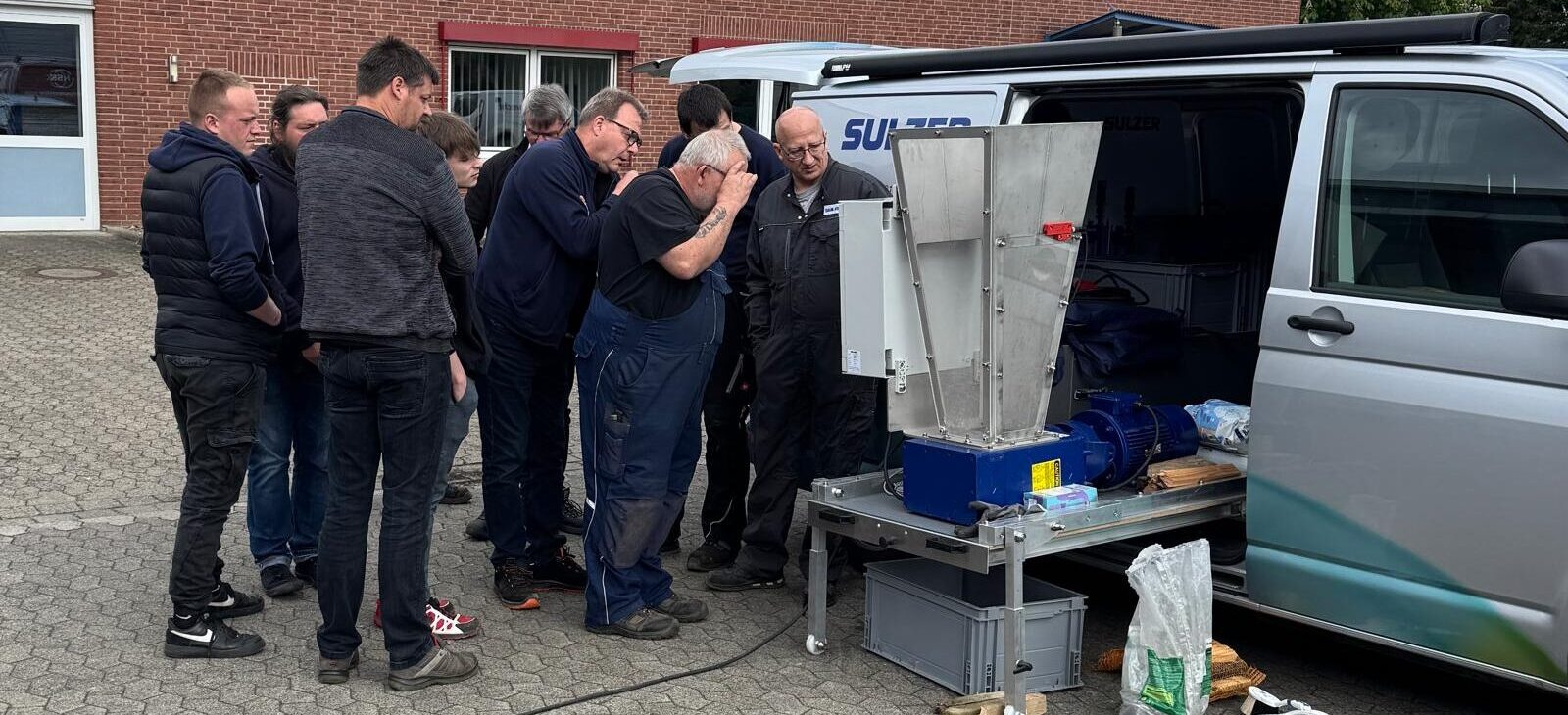 Mehrere Personen stehen dicht beieinander und betrachten eine offene Maschine neben einem Transporter auf einem gepflasterten Hof während der Schulung.