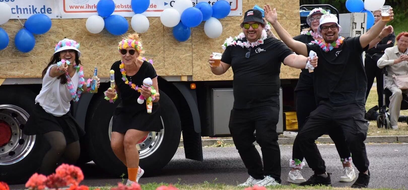 Vier Mitarbeiter von Ihrke mit Blumenketten und Getränken tanzen vor einem festlich geschmückten Umzugswagen mit Luftballons und Werbebanner auf dem Malchower Volksfest.