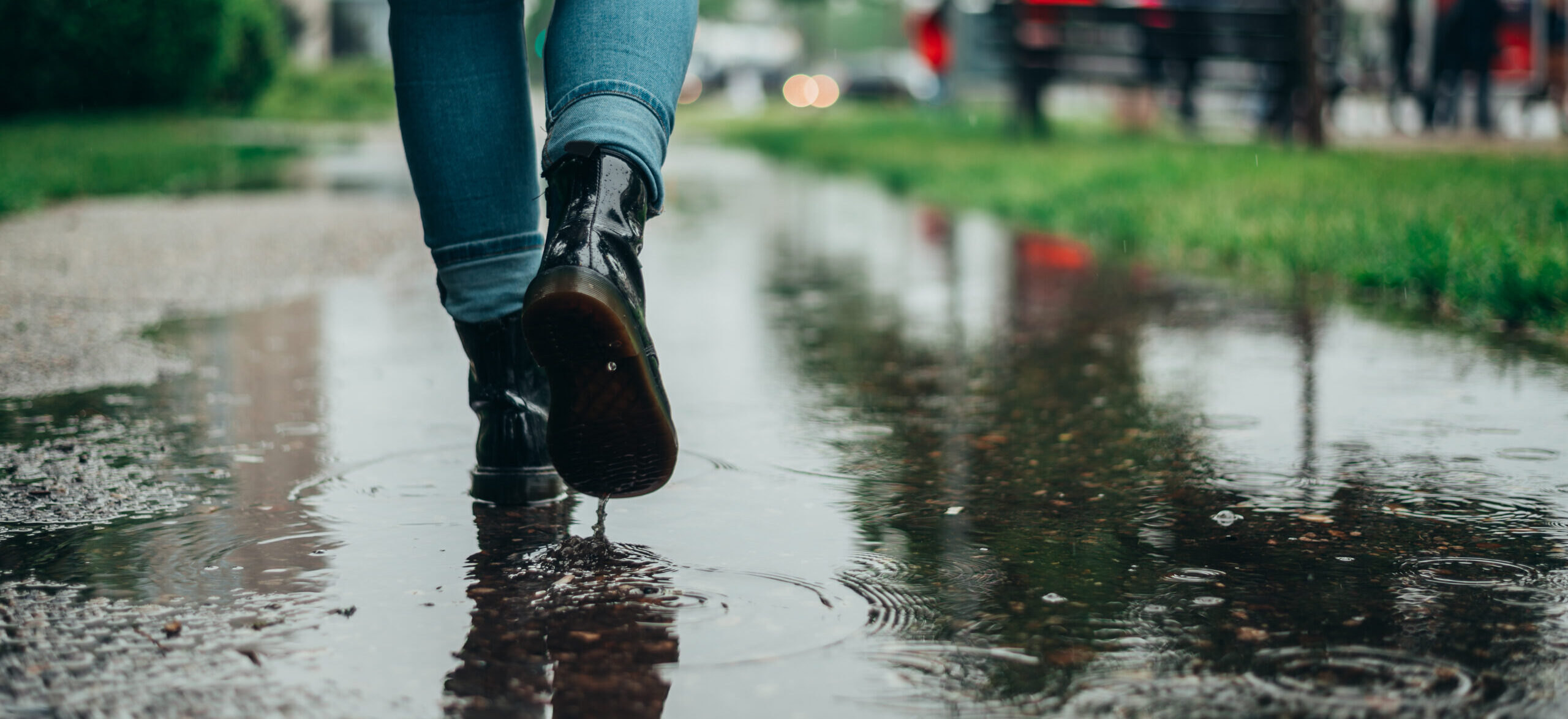 Frau mit Mantel und Stiefeln läuft durch eine große Pfütze auf einem Gehweg, während es regnet, was auf den Einsatz einer Regenwasserpumpe hinweist.