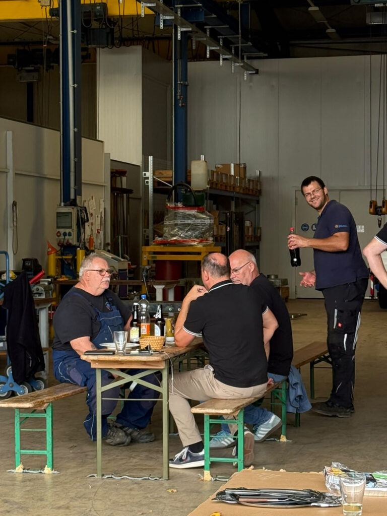 Mehrere Männer sitzen an einem Tisch in einer Werkhalle, einer steht daneben und hält eine Flasche.