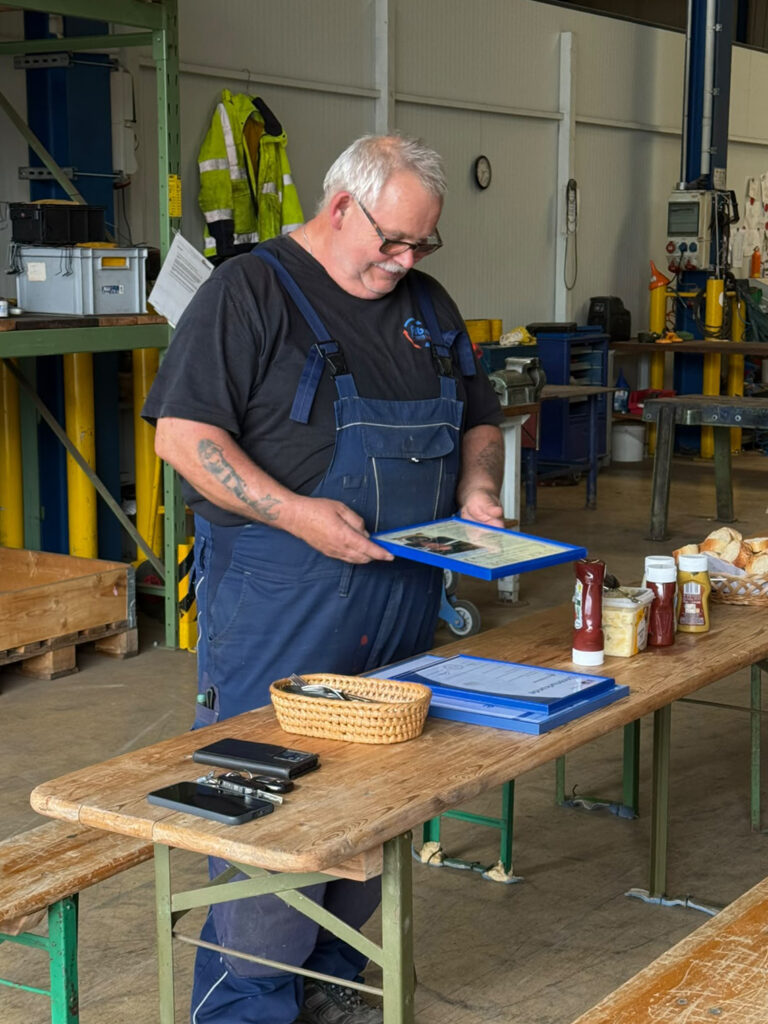 Mann mit Brille und blauen Latzhosen steht an einem Tisch mit Tabletts, Brötchen, Ketchup- und Senfflaschen in einer Werkhalle. Er häjt eine Urkunde für seine 40 Jahre bei Albrecht Maschinenbau in den Händen.