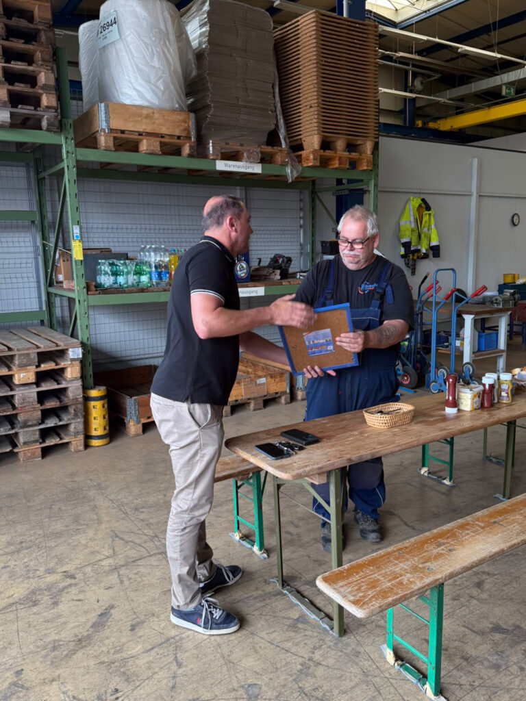 Zwei Männer stehen an einem Holztisch in einer Lagerhalle und halten gemeinsam ein Dokument.
